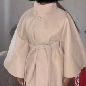 Cream poncho med bälte och vida ärmar - Säljer en beige poncho med snyggt bälte i midjan och vida ärmar. Jackan har hög krage och en dekorativ knapp upptill. Perfekt för dig som vill ha en stilren och trendig look under kallare dagar. 