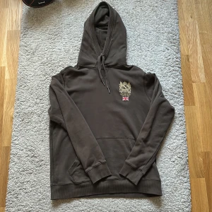 Brun hoodie från Morris, storlek M - Säljer en brun hoodie från Morris med snyggt broderat emblem och brittisk flagga på bröstet. Tröjan har huva med snörning, känguruficka och ribbade muddar. Perfekt för chill dagar och enkel att matcha med jeans eller joggers.
