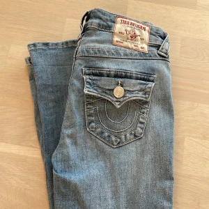 True Religion ljusblå jeans med detaljer 25W - Säljer ett par ljusblå jeans från True Religion med snygga kontrastsömmar och ikonisk bakficka med lock och silverfärgad knapp. Klassisk femficksmodell i mjuk bomullsdenim, perfekt för dig som gillar street och premiumkänsla.