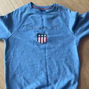 Blå sweatshirt från GANT med emblem - Blå sweatshirt från GANT i storlek XS med broderat GANT-logo och ett snyggt emblem med stjärnor och ränder på bröstet. Tröjan har rund hals och korta ärmar, tillverkad i mjuk bomull. Perfekt för dig som gillar klassisk och sportig stil.