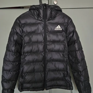 Svart dunjacka från Adidas, stl S - Säljer en svart dunjacka från Adidas i storlek S. Jackan har huva, dragkedja framtill och klassisk Adidas-logga på bröstet. Den är quiltad och har en clean, sportig look som passar perfekt när det är kallt ute.