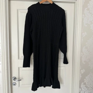 Svart stickad klänning från Vero Moda AWARE - Supersnygg svart stickad klänning från AWARE med kabelstickat mönster och volangkant nedtill. Klänningen har lång ärm, rund hals och markerad midja som ger en schysst siluett. Perfekt höstklänning. Oanvänd, endast provad 