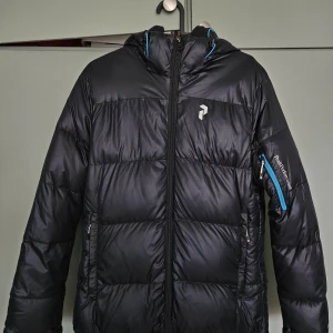 Svart pufferjacka Peak Performance S - Säljer en svart pufferjacka från Peak Performance i storlek S. Jackan har huva, blå detaljer vid dragkedjor och logga på bröstet samt ärmen. Perfekt för kalla dagar och har en sportig vibe. Flera fickor med dragkedja och riktigt skön passform.