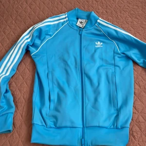 Blå Adidas track jacket med vita ränder - Säljer en klassisk blå Adidas kofta med vita ränder längs ärmarna och vit logga på bröstet. Koftan har dragkedja framtill, ståkrage och två fickor. Tillverkad i polyester och har en sportig vibe som passar perfekt till streetwear.