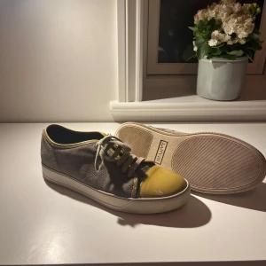 Lanvin sneakers i mocka och lack - Snygga sneakers från Lanvin i grå mocka med gul lackad tå och gul kant runt öppningen. Klassisk låg modell med vita snören och vit sula. Perfekt för dig som vill sticka ut med en unik färgkombo och exklusiv känsla. Priset är ej hugget i sten!