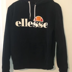 Svart Ellesse hoodie med logga - Svart hoodie från Ellesse med stor vit logga och färgglad detalj på bröstet. Tröjan har huva med vita snören och en klassisk känguruficka framtill. Materialet är mjukt och består av 80% bomull och 20% polyester. Perfekt för chill dagar eller när du vill ha en sportig look.