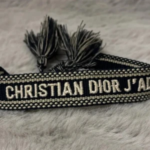 Christian Dior J'adior armband - Pris går att diskutera.Snyggt svart armband från Christian Dior med broderad text 'J'ADIOR' i beige. Armbandet har dekorativa tofsar i ändarna och ett unikt, trendigt utseende som sticker ut. Perfekt accessoar för att lyfta din outfit.