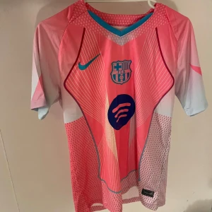 Barcelona rosa fotbollströja Nike S - Barcelona fotbollströja i en fräsch rosa färg med ljusblå detaljer och vita inslag. Tröjan har korta ärmar, tunna vita linjer och prickigt mönster på sidorna. FCB-logga och Spotify-tryck på bröstet, samt Nike swoosh. Tillverkad i lätt polyester för max komfort.