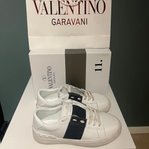 Valentino Garavani Open sneakers vit/blå - Snygga Valentino Garavani Open sneakers i vitt skinn med bred mörkblå rem över vristen och coola nitar på sulan. Klassisk rund tå och platt sula, perfekta för en trendig streetstyle-look.