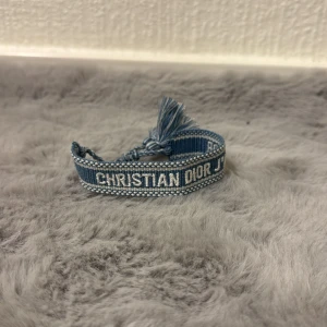 Blått armband från Christian Dior - Snyggt blått armband från Christian Dior med vit broderad logga och tofsdetaljer. Armbandet är gjort i textil och har en cool, avslappnad vibe som passar perfekt till jeans eller som en statement-accessoar.