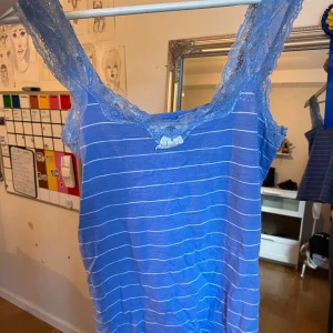 Blå randig linnetopp från Hunkemöller - Supersöt blå linnetopp från Hunkemöller med tunna vita ränder och breda spetsband. Toppen har en liten rosett framtill och v-ringad hals. Perfekt för chill dagar eller när du vill känna dig extra comfy. Materialet är mjukt och lätt, med fina spetsdetaljer Den är så otroligt bekväm och så snygg till ALLT☺️💕 