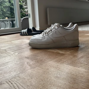 Vita Nike Air Force 1 sneakers - Klassiska vita Nike Air Force 1 sneakers med lågt skaft och ikonisk swoosh på sidan. Skorna har snörning, perforerad tå och är tillverkade i skinn. Perfekta för en clean och stilren look.