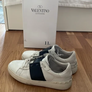 Valentino Garavani vita sneakers - Snygga vita Valentino Garavani sneakers med bred svart detalj över sidan. Klassisk rund tå, platt sula och snörning. Tillverkade i skinn för en lyxig känsla och stilren look. Perfekta för dig som vill sticka ut med designerdojor.