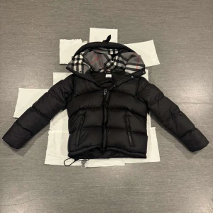 Svart pufferjacka från Burberry - Säljer en svart pufferjacka från Burberry med avtagbara ärmar och huva fodrad i klassiskt rutigt tyg. Jackan har dragkedja, två fickor med dragkedja och justerbar dragsko nertill. Perfekt för kalla dagar och kan bäras både som jacka och väst.