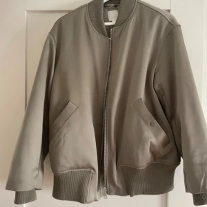 Beige bomberjacka med dragkedja - Snygg beige bomberjacka med ribbade muddar och krage. Jackan har två fickor med knappstängning och en stilren dragkedja framtill. Perfekt för dig som gillar en clean och enkel look. Passformen är oversized och materialet känns mjukt och slitstarkt.