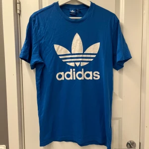 Blå Adidas t-shirt med stor logga - Blå t-shirt från Adidas med stor vit Trefoil-logga och text på bröstet. Klassisk rund hals och korta ärmar. Tillverkad i mjuk bomull, perfekt för en sportig och avslappnad stil.