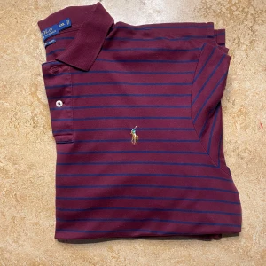 Randig vinröd pikétröja Polo Ralph Lauren - Snygg vinröd pikétröja från Polo Ralph Lauren i storlek XXL. Tröjan har tunna marinblå ränder, klassisk krage och broderad logga på bröstet. Tillverkad i mjuk bomull (Pima Soft Touch) och har korta ärmar. Perfekt för en chill och stilren look.