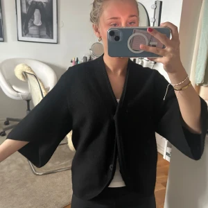 Svart kofta från Dagmar med vida ärmar - Svart kofta från Dagmar med oversized passform och vida trekvartsärmar. Koftan har v-ringad öppning med knappar, och elastisk detalj i ryggen som ger en snygg siluett. Tillverkad av ull. 
