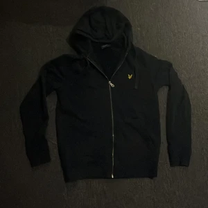 Svart hoodie från Lyle & Scott - Mörkblå hoodie från Lyle & Scott med dragkedja och huva. Klassisk gul logga på bröstet och långa ärmar. Perfekt för chill dagar eller när du vill ha en enkel och stilren look. Gjord i mjuk bomull för extra komfort. Skriv till mig om du undrar över nått.PRISET KAN DISKUTERAS 