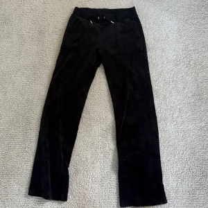 Svarta mjukisbyxor Juicy Couture S - Svarta mjukisbyxor från Juicy Couture i storlek S. Byxorna har bred resår i midjan med snörning, stora fickor fram och bak med knappdetaljer. Mjukt och lyxigt material som känns skönt mot huden. Perfekt för chill dagar eller när du vill vara extra comfy.