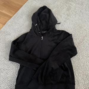 Svart Juicy Couture hoodie i velour - Svart hoodie från Juicy Couture i mjuk velour med dragkedja och huva. Klassisk J-dragkedja och snörning i luvan. Perfekt för en chill och trendig look. Långärmad och riktigt skön att ha på sig.