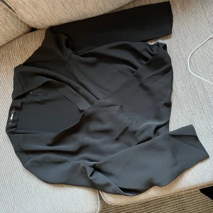Svart omlottblus från Bikbok  - Stilren svart omlottblus från Bikbok med v-ringning och långa ärmar. Blusen har en mjuk och flowy känsla, perfekt för att lyfta vilken outfit som helst. Enkel att matcha och ger en snygg siluett.