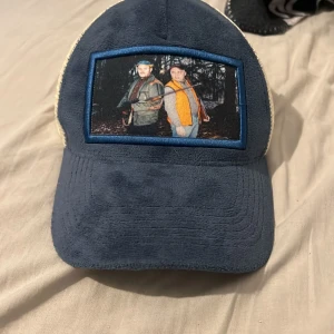 Leif o Billy keps - Säljer en unik truckerkeps från LL CAPS med blå manchester framtill och beige mesh baktill. Framsidan har en broderad patch med ett foto på två personer i skogen. Perfekt för dig som vill sticka ut med en personlig stil.