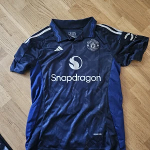 Manchester United Mainoo #37 bortatröja - Snygg Manchester United bortatröja med Mainoo och nummer 37 på ryggen. Tröjan är marinblå med vita detaljer, har korta ärmar och krage. Tillverkad i lätt och ventilerande polyester med diskret mönster. Officiell Snapdragon sponsorlogga på bröstet.