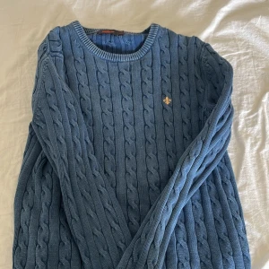 Blå stickad tröja från Morris - Säljer en blå kabelstickad tröja från Morris med rund hals och lång ärm. Tröjan har ett broderat Morris-emblem i gult på bröstet och är gjord i mjuk bomull. Perfekt för dig som gillar klassisk stil med en modern twist.