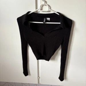 Svart ribbad croppad topp H&M Divided - Svart långärmad croppad topp från H&M Divided i ribbad bomull. Snygg v-ringad design med krage som ger en cool och trendig look. Perfekt att matcha med höga jeans eller kjol för en stilren vibe.