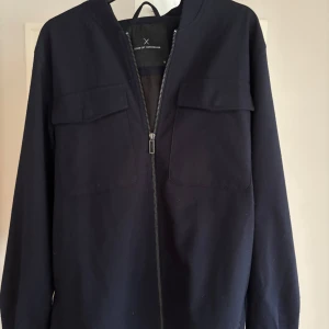 Mörkblå overshirt från Clean Cut Copenhagen - Snygg mörkblå overshirt från Clean Cut Copenhagen med dragkedja framtill och två stora bröstfickor. Jackan har långa ärmar med tryckknappar vid mudden och är tillverkad i ett mjukt material som känns riktigt skönt. Perfekt för lager-på-lager-stil. Är i nyskick. Bara att skriva vid frågor eller funderingar!