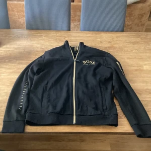 Svart ziptröja från Hugo Boss M - Svart långärmad ziptröja från Hugo Boss med gulddetaljer och logga på bröstet samt på kragen. Tröjan har hel dragkedja framtill och ribbade muddar. Snygg och stilren modell som passar perfekt till streetwear eller chill dagar.