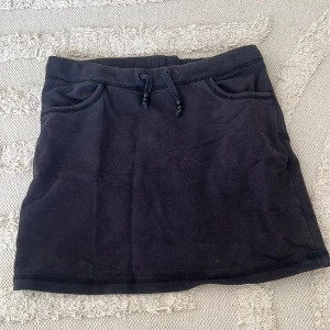 Svart kortkjol från Marwin - Svart kortkjol från Marwin i mjuk bomull. Kjolen har en enkel design med två fickor fram och snörning i midjan för extra chill vibe. Perfekt för dig som gillar basic och comfy stil. Passformen är normal och materialet känns skönt mot huden.