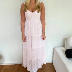 Ljusrosa långklänning med volanger - Säljer en superfin ljusrosa långklänning med volanger i flera lager. Klänningen har v-ringad hals, knappar framtill och är ärmlös. Endast använd en gång.