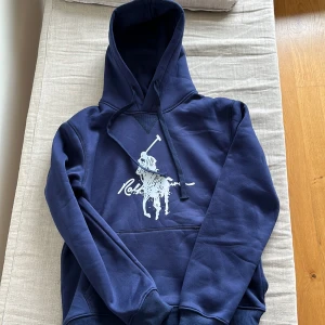 Marinblå hoodie från Ralph Lauren - Snygg marinblå hoodie från Ralph Lauren med stor vit Polo-logga på bröstet. Tröjan är i väldigt bra skick och perfekt till hösten. Storleken är L men skulle även säga att den passar M. Hör av er vid frågor!