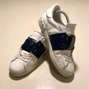 Valentino Garavani Open sneakers vit/blå - Snygga Valentino Garavani Open sneakers i vitt skinn med bred marinblå läderdetalj över mitten. Klassisk rund tå, vita snören och coola nitar på sulan. Perfekta för dig som gillar exklusiva och stilrena sneakers med en twist. Dustbag ingår    Här du frågor är det bara att höra av sig 