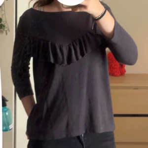 Mörkgrå tröja med volangdetalj - Säljer en mörkgrå tröja med snygg volangdetalj över bröstet. Tröjan har trekvartsärm och en avslappnad passform, perfekt för dig som gillar en enkel men ändå trendig look. Materialet känns mjukt och skönt mot huden.