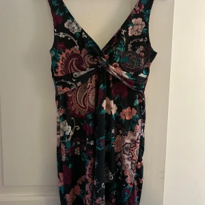 Blommig långklänning från Gina Tricot - Supersnygg långklänning från Gina Tricot med blommigt mönster i vinrött, rosa, beige och grönt på svart botten. Klänningen har omlott-topp med twistad detalj framtill och breda axelband. Perfekt för dig som vill sticka ut med färg och mönster!