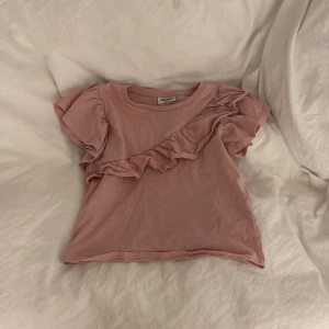 Rosa volangtopp från Kappahl - Supersöt rosa topp från Kappahl med volangdetalj snett över framsidan och puffiga korta ärmar. Tillverkad i mjuk bomull, perfekt för dig som gillar gulliga och lekfulla plagg. Enkel att matcha med jeans eller kjol för en chill look.