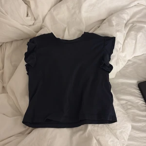 Svart volangtopp från Gina Tricot - En svart topp från Gina Tricot med korta ärmar och snygga volangdetaljer på axlarna. Tillverkad i mjuk bomull, perfekt för dig som gillar en enkel men ändå trendig look. Passar till allt och är superenkel att styla.