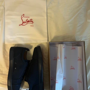 Mörkblå sneakers från Christian Louboutin - Snygga mörkblå sneakers från Christian Louboutin i mocka med klassisk röd sula och diskret broderad logga på hälen. Skorna har rund tå, snörning och en stilren design som sticker ut med den ikoniska röda undersidan.