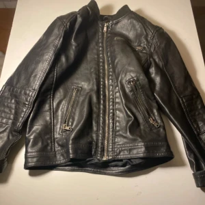 Svart skinnjacka från H&M - Snygg svart skinnjacka från H&M med coola detaljer som dragkedjor på fickorna och framtill. Jackan har en klassisk bikerstil med ribbad krage och långa ärmar.