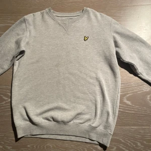 Grå sweatshirt från Lyle & Scott - Säljer en stilren grå sweatshirt från Lyle & Scott Junior med klassisk rund hals och broderad gul logga på bröstet. Tröjan har ribbade muddar vid ärmslut och nederkant, perfekt för chill dagar. Materialet är mjuk bomull som känns skönt mot huden.