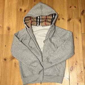 Grå Burberry hoodie med dragkedja - Snygg grå hoodie från Burberry med klassiskt rutigt foder i huvan och broderad logga på bröstet. Tröjan har dragkedja, snörning i huvan och två fickor framtill. Perfekt för dig som gillar stilren streetwear med lyxig touch.