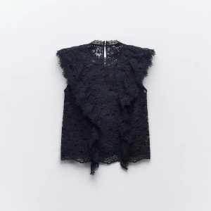 Svart spetsblus med volang - Supersnygg svart blus i spets med volangdetaljer framtill och ärmlös design. Blusen har rund halsringning och knäppning bak i nacken. Perfekt för dig som gillar romantiska och detaljerade plagg.