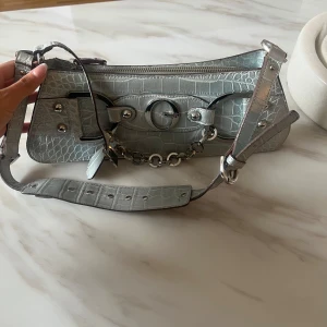 Silver axelväska från Guess med kedjedetalj - Säljer en snygg silverfärgad axelväska från Guess i croco-mönstrat skinnimitation. Väskan har justerbart axelband, coola metalldetaljer och kedja framtill. Invändigt finns ett foder med Guess-logga och dragkedjefack. Perfekt för dig som gillar statement-accessoarer.