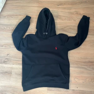 Svart hoodie från Ralph Lauren - Svart hoodie från Ralph Lauren med klassisk röd broderad logga på bröstet. Tröjan har huva med snörning, känguruficka och ribbade muddar. Perfekt för dig som gillar en clean och stilren look.