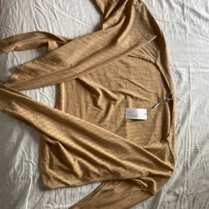 Beige ribbad omlottkofta från NLY Trend - Snygg beige omlottkofta från NLY Trend i ribbad bomullsmix. Koftan har långa ärmar och knytband i midjan för en trendig look. Perfekt att slänga över en topp eller klänning för extra stilpoäng.