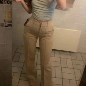 Beiga kostymbyxor från NA-KD, strl 36 - Snygga beiga kostymbyxor från NA-KD i storlek 36. Byxorna har hög midja, raka ben med lätt bootcut och pressveck framtill. Perfekta för en clean och stilren look. Tillverkade i ett mjukt material som sitter bekvämt hela dagen. Aldrig använd. Utan prislapp