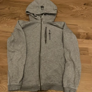 Grå hoodie från Seal Racing - Säljer en grå hoodie från Seal Racing med dragkedja framtill och en extra zip på bröstet. Tröjan har huva med tryckt logga och långa ärmar. Perfekt för chill dagar och har en clean look som funkar till jeans eller joggers.